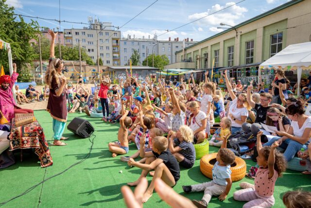 Lato na Madalinie - dzieci siedzące na zielonym podłożu, wyciągające w górę ręce